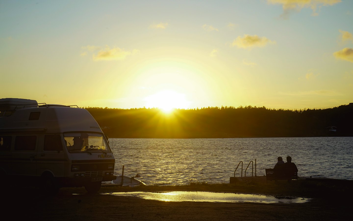 To personer sitter på en benk og ser på solnedgangen ved en bobil.