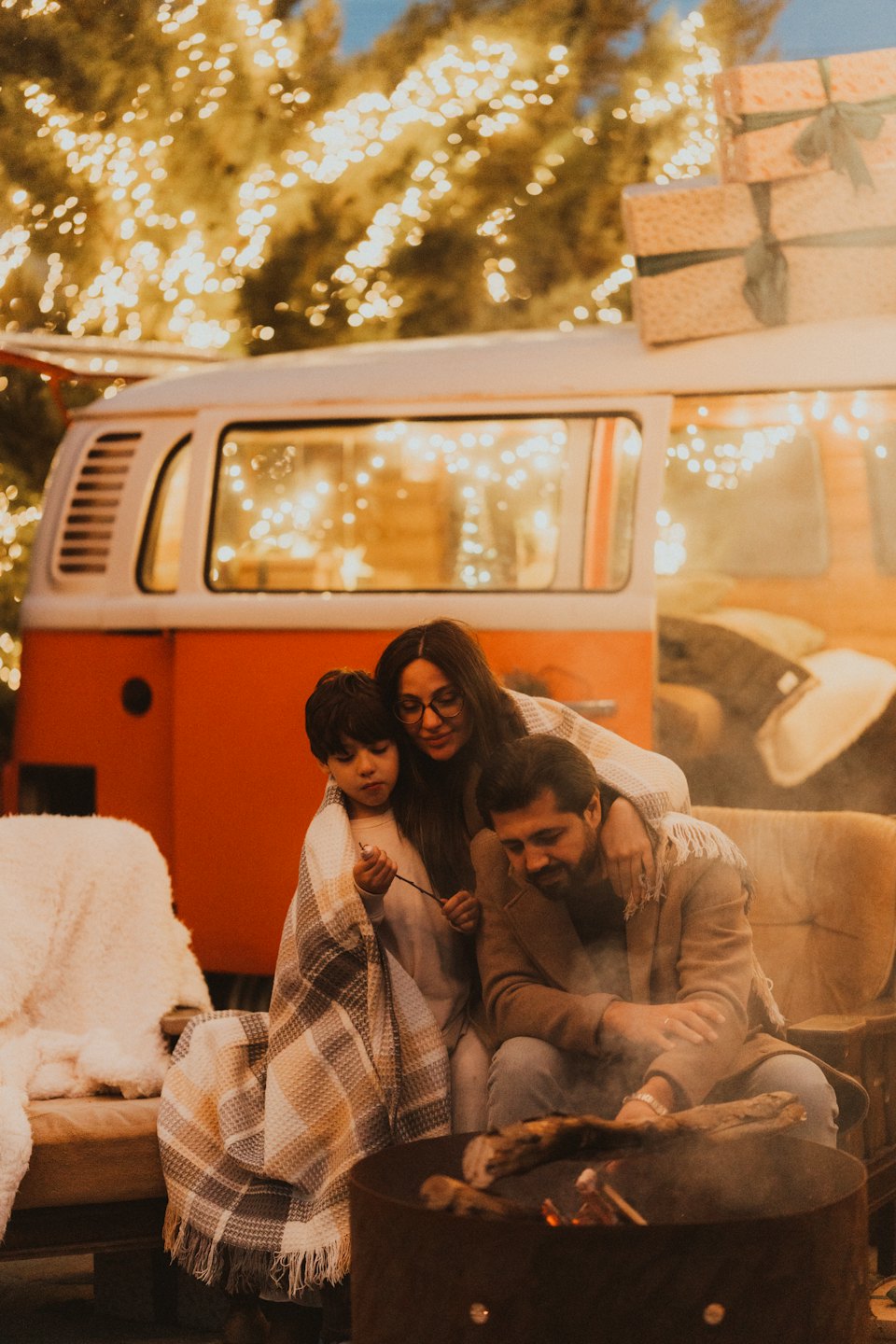 Familie samles under et teppe ved et bål nær campingvognen deres.